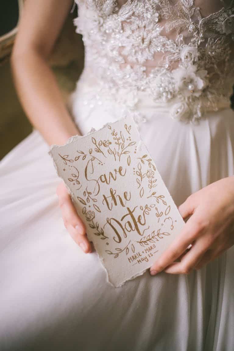 A close-up of a bride holding an elegant 'Save the Date' card with intricate calligraphy.