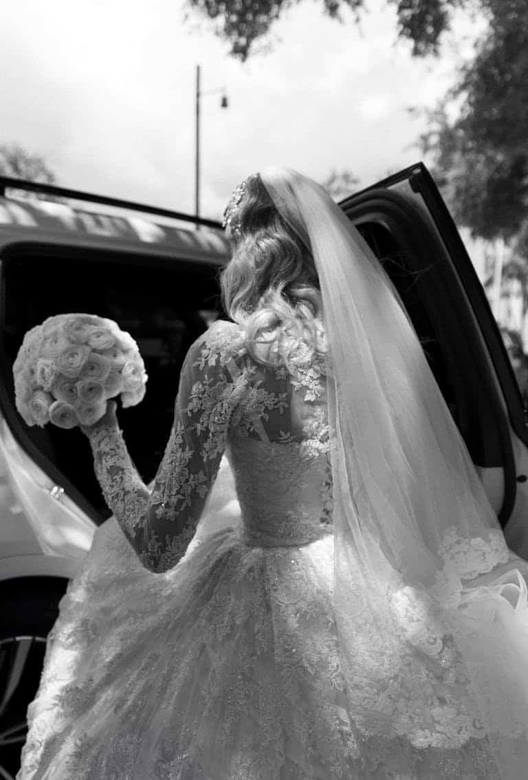 Bride in lace dress with bouquet entering car, elegant and classic moment.